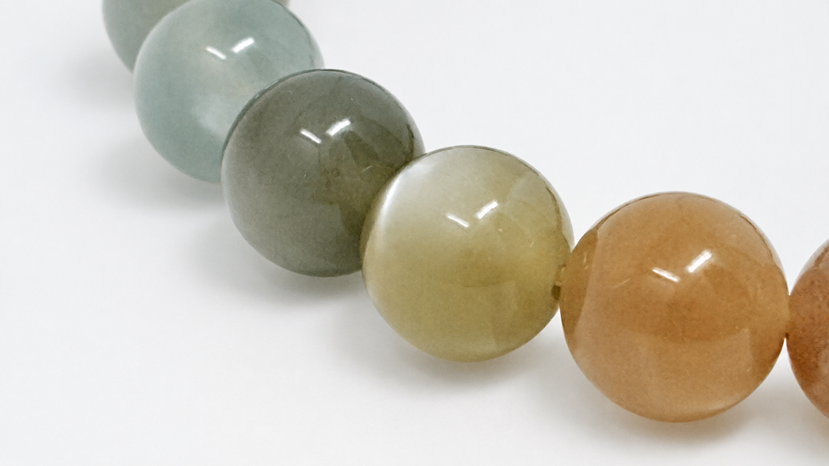 A close-up of polished round gemstone beads in a row, showing translucent shades of pale green, gray-green, yellow-green, peachy orange, and a small portion of a darker brown bead on a white background.